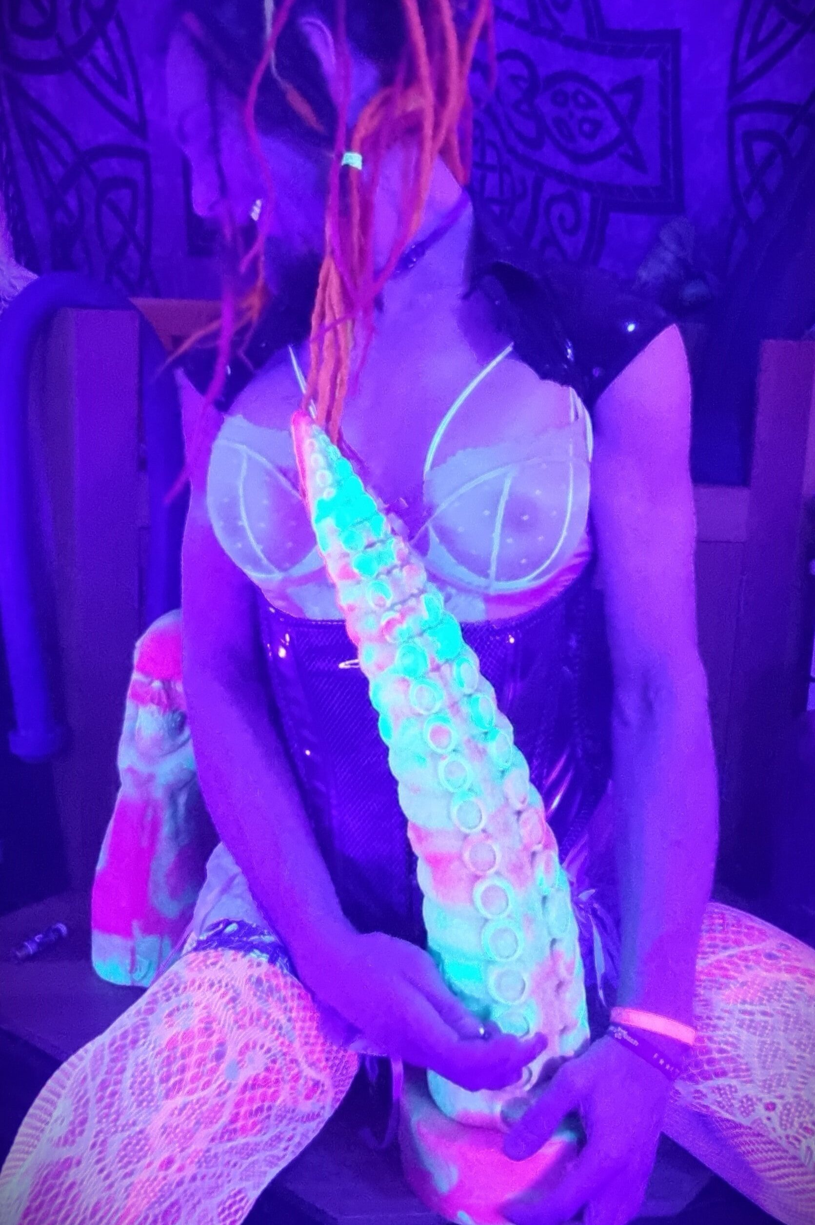 Jasmine Dlight posing in UV light with rainbow tentacle dildo, wearing sheer lingerie and PVC corset.