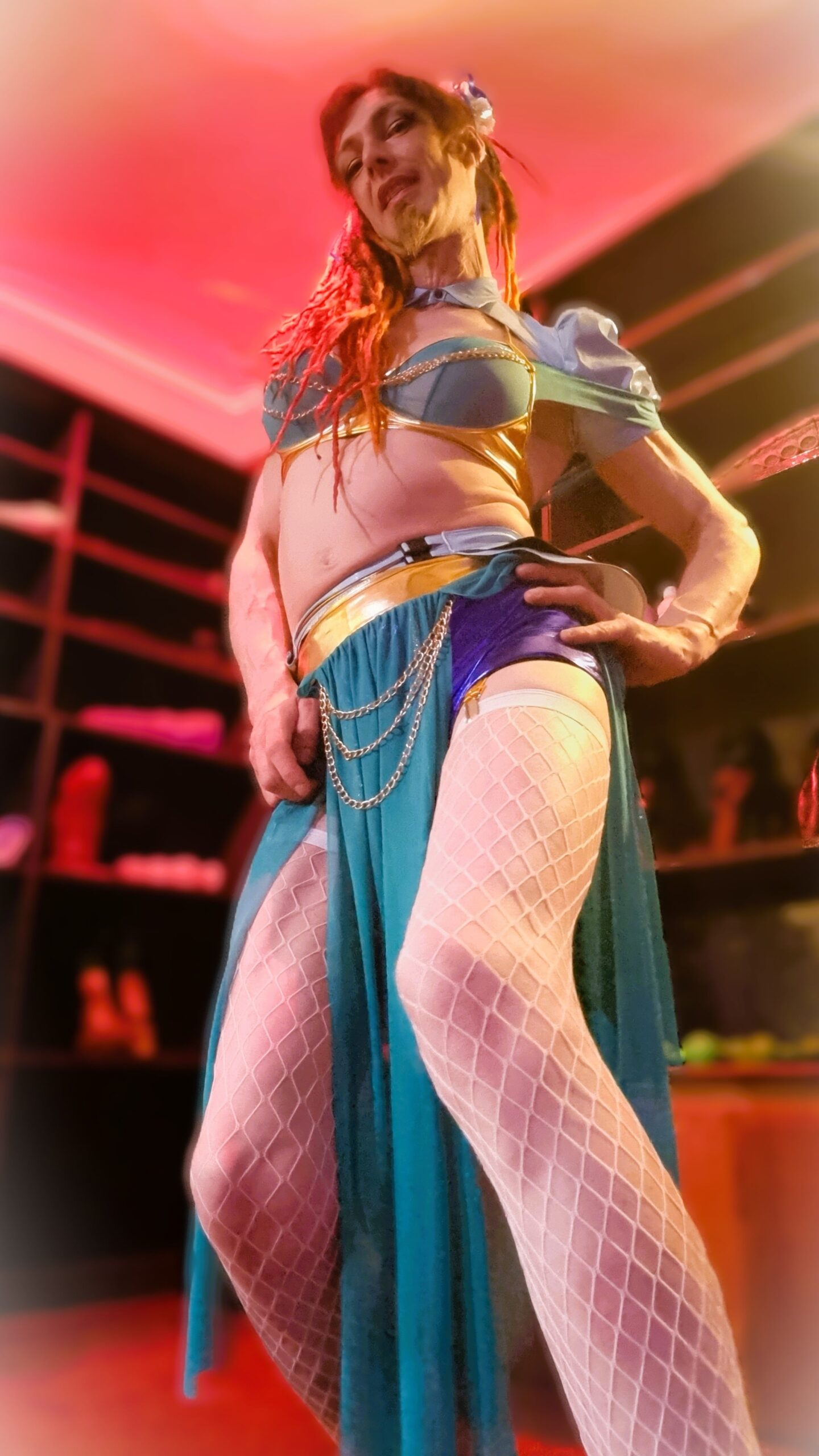 Jasmine_Dlight dressed as a seductive fantasy princess in shimmering teal and gold, posing confidently in front of a glowing wall of toys at House Edonistica.